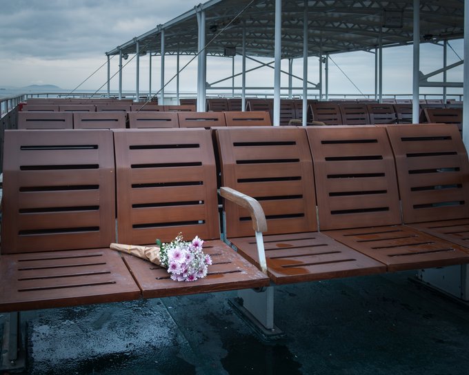 An empty row of wooden slatted deck chairs on the deck of a ferry or boat, with a small bouquet of pink and white flowers wrapped in paper resting on one seat against a backdrop of cloudy skies and distant land.