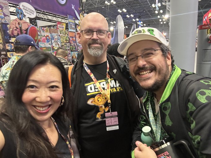 A selfie of three smiling people at what appears to be a comic convention, including one wearing a 'Peeps vs. Marshmallows' t-shirt.