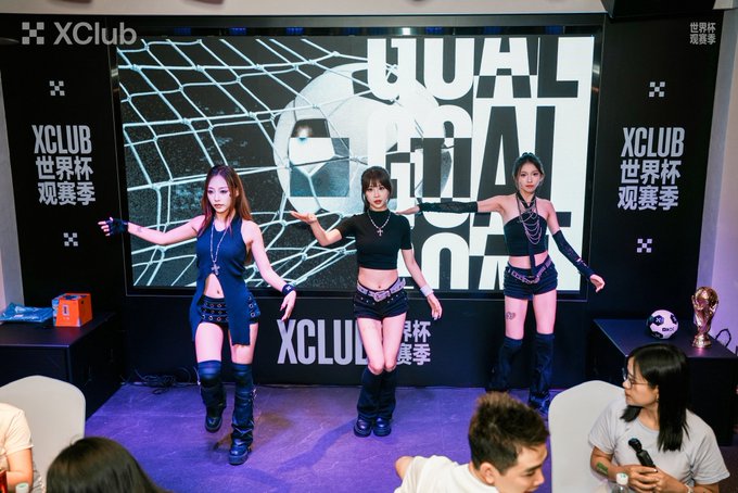 Three women dancing at an XClub World Cup viewing event ('世界杯观赛季') with a soccer goal backdrop, while spectators watch.