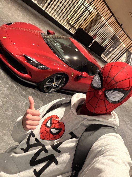 A person wearing a Spider-Man mask and a white Spider-Man hoodie gives a thumbs-up in front of a red Ferrari sports car.