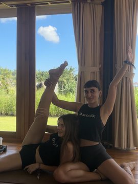 Two young women pose together during a flexibility/stretching session in Bali, with one in a black "BROOKS" top and the other in matching black athletic wear, as the background shows tropical greenery through floor-to-ceiling windows. The image ties into the post's promotion of strcUSX, an accessible onchain yield product that—as the caption notes—requires no DeFi expertise, just a simple deposit to start earning.
