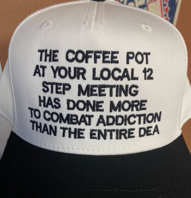 A white and black baseball cap has embroidered text reading: 'THE COFFEE POT AT YOUR LOCAL 12 STEP MEETING HAS DONE MORE TO COMBAT ADDICTION THAN THE ENTIRE DEA'.