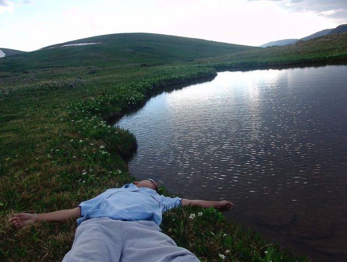 A man in a light blue shirt and gray pants lies peacefully on a grassy shoreline beside a tranquil lake, with his arms outstretched as if embracing the serene landscape. The image evokes a sense of calm and contentment, reflecting the relaxed, optimistic vibe of the "Gm frens of Rex Wagmi" message.