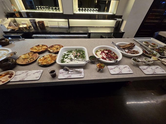 A buffet counter displays an assortment of dishes including salads, meats, and sauces in serving bowls and trays, with empty marble-patterned plates ready for diners.