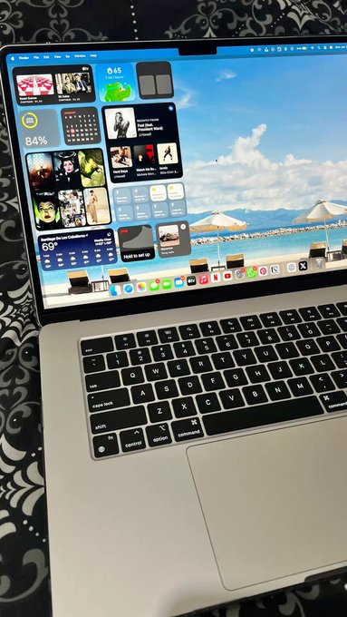 An open silver MacBook Pro laptop displays a macOS desktop with widgets including weather, calendar, and music, set against a beach wallpaper.