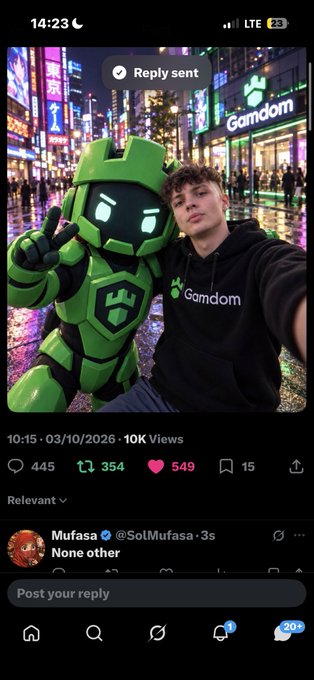 A young man in a black 'Gamdom' hoodie takes a selfie with a green robotic mascot in a vibrant, neon-lit city street at night, with a Twitter post visible showing 10K views, 445 comments, and a reply from 'Mufasa' saying 'None other'.