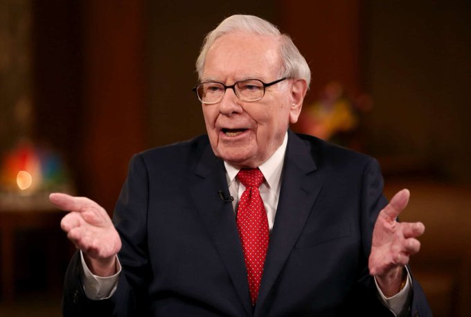 An elderly man wearing glasses, a dark suit, and a red polka dot tie gestures with both hands while speaking, appearing to be in the middle of an interview.