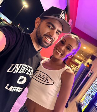 A selfie of a smiling man and woman; the man wears a black 'CONFIRMED LAS VEGAS' t-shirt and an LA Dodgers cap, while the woman wears a white 'Von Dutch' crop top.