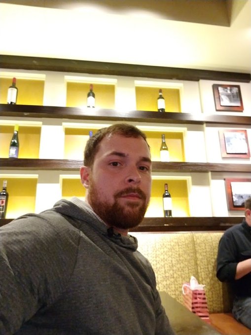 A selfie of a bearded man in a gray hoodie sitting in a restaurant booth, with shelves displaying wine bottles and framed pictures behind him.