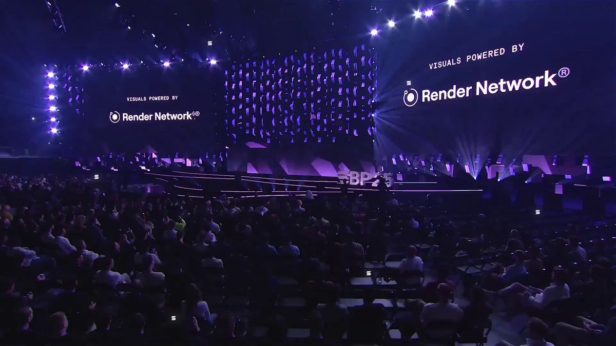 The image depicts a crowded auditorium filled with people, standing near rows of chairs arranged throughout the space. There are speakers placed on both sides of the auditorium facing the audience. The scene also features several screens displaying information about the event and its sponsors. The main focus in this image is a large screen displaying an advertisement for " Render Network".