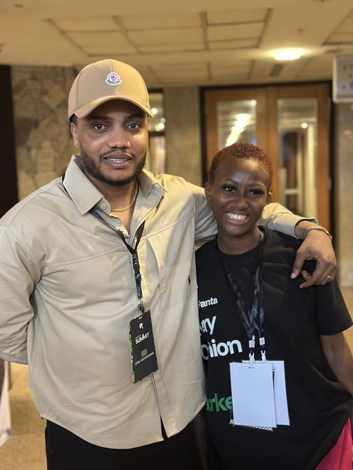 A photograph of two smiling people posing together indoors; the man on the left wears a cap and tan shirt with a 'STAFF' lanyard, while the woman on the right wears a black shirt with text mentioning 'Atlanta' and 'Action'.