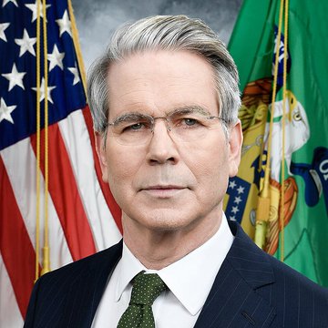 An official portrait of a man in a dark suit with a green tie, wearing glasses, posed in front of an American flag and a green flag with a seal.