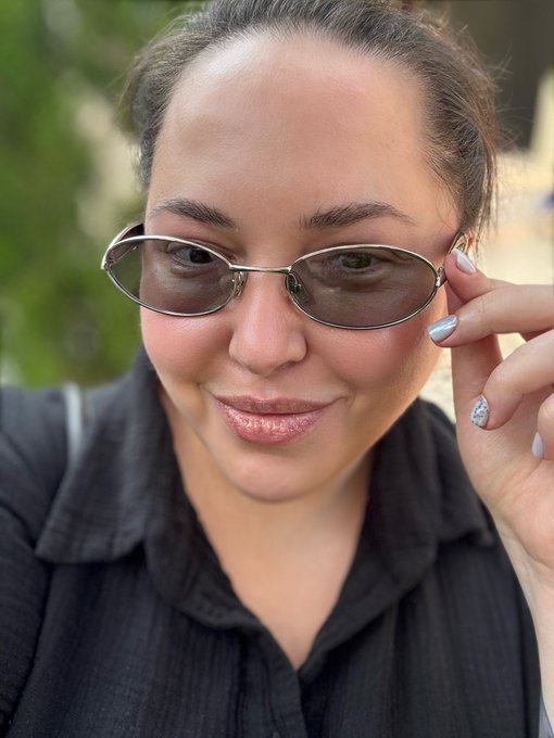 A close-up selfie of a smiling woman with dark hair pulled back, wearing small oval sunglasses and a black collared shirt, with sparkly silver nail polish.