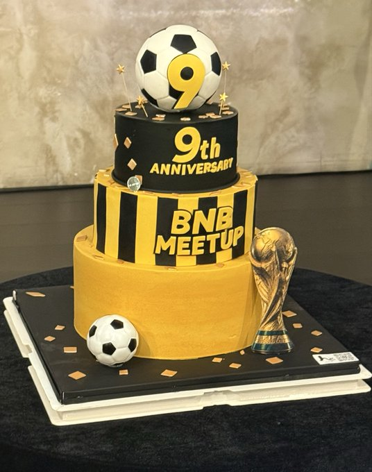 A custom tiered yellow and black birthday cake celebrating a '9th ANNIVERSART BNB MEETUP', decorated with a soccer ball bearing a '9', a World Cup trophy, and a smaller soccer ball.