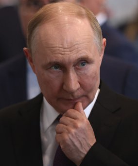 A close-up portrait of Vladimir Putin in a dark suit and tie, posing thoughtfully with his hand on his chin against a blurred background.