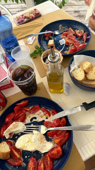 An overhead view of a dining table set with a meal of fresh tomatoes, mozzarella cheese, and bread, accompanied by a glass of dark soda, a bottle of olive oil, and a bottle of water.