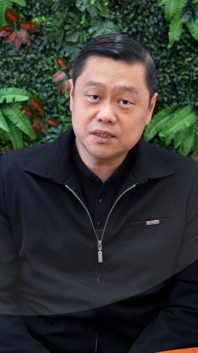 A portrait of an East Asian man with short black hair wearing a black zip-up jacket, sitting in front of a green leafy plant wall background.