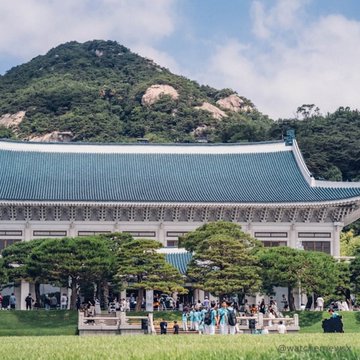 The image shows the Blue House (Cheong Wa Dae) in Seoul, South Korea, the former executive office and official residence of the South Korean president, set against a backdrop of lush mountains and framed by manicured pine trees, with visitors gathered at its entrance. The scene visually anchors the accompanying news about South Korea piloting tokenized government bonds on the Bank of Korea's wholesale CBDC rail in 2027—symbolizing how the nation is modernizing its traditional financial and governmental infrastructure ("legacy plumbing") with new tokenized securities rules.