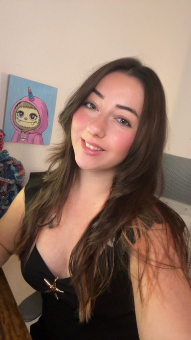A smiling woman in a black top takes a selfie indoors, with a cartoon painting of a unicorn character visible on the wall behind her.