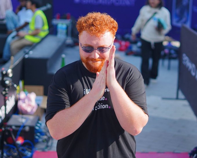 A red-haired, bearded man in sunglasses stands with his hands pressed together in a grateful gesture, smiling at the camera at the Solana Summit CA event in Canada, where he is set to speak about distribution-maxxing and related topics.