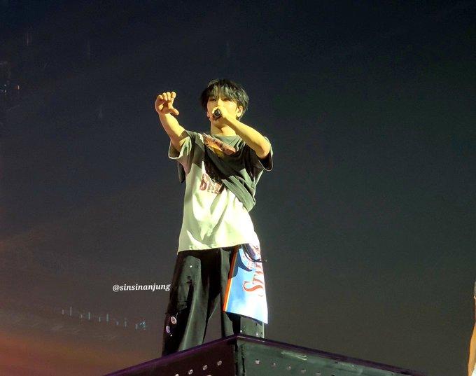 Concert photo of a young male performer holding a microphone on stage, wearing a white t-shirt and graphic shorts, watermarked @sinsinanjung.