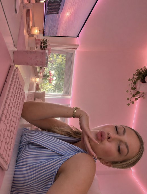 A selfie of a young woman with blonde hair in a pink room, wearing a blue striped halter top with her hand resting near her face.