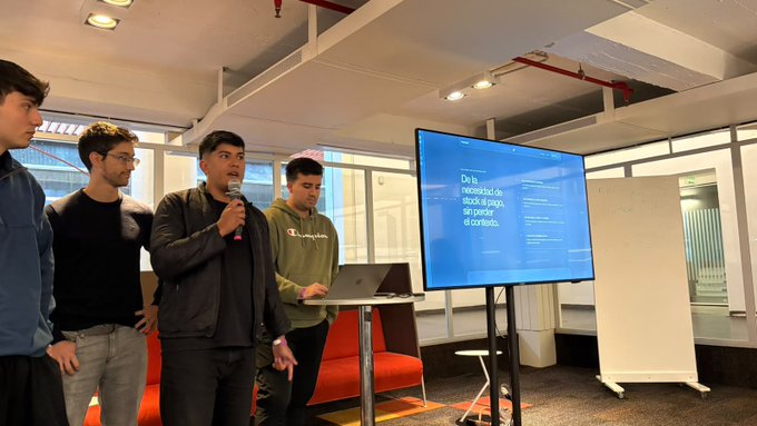 A group of young men giving a presentation in a modern office space, with one speaking into a microphone next to a screen displaying 'De la necesidad de stack al pago, sin perder el contexto.'