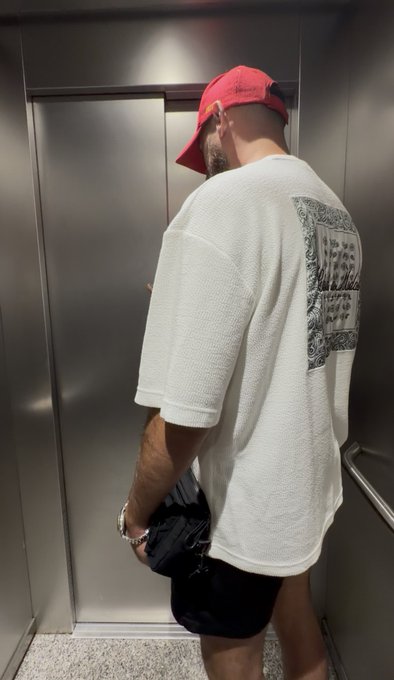 A man wearing a red backwards cap and an oversized white t-shirt with a paisley bandana print on the back stands in an elevator holding a black bag. The "Gm friends" greeting pairs with the casual morning-ready streetwear vibe of the relaxed fit tee and cap.