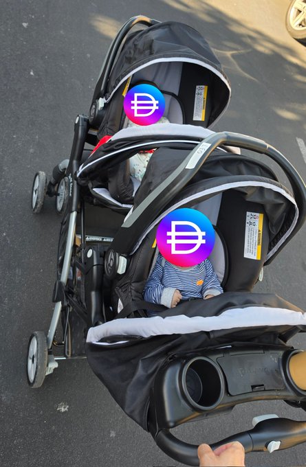 The image shows two babies sitting in double strollers with black seats and white wheels. The stroller handles are attached to the front of the stroller, allowing for easy maneuvering. A person's hand can be seen holding onto one of the handles while another handle is visible on the side of the stroller. In the background, there are some cars parked along a street.