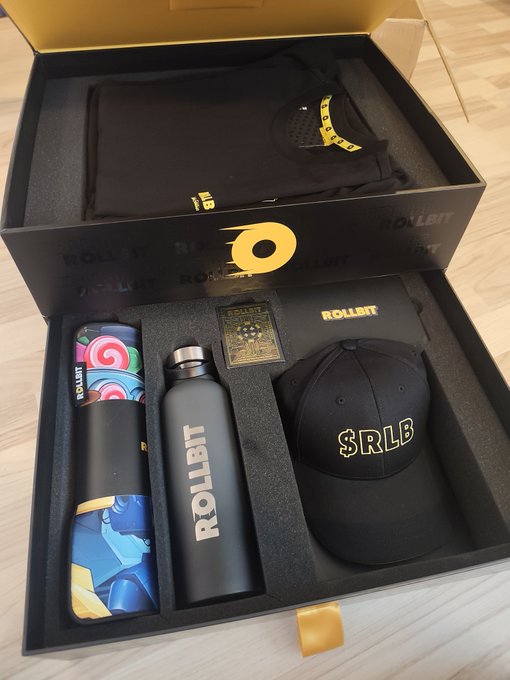 An open Rollbit-branded lootbox reveals a premium merch package featuring a black "$RLB" cap, a matte black Rollbit water bottle, a hoodie with a face mask, a Rollbit pouch, and a trading card, all arranged neatly in custom foam packaging. The items prominently display the "ROLLBIT" branding and the "$RLB" ticker, tying directly to the celebratory unboxing post.