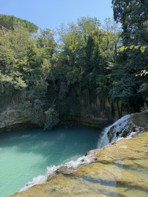 A serene natural pool with turquoise water sits beneath a small cascading waterfall, surrounded by lush green trees and rocky cliffs under a clear blue sky. The tranquil landscape perfectly captures the peaceful "Good Morning" spirit of a fresh, sunlit start to the day.