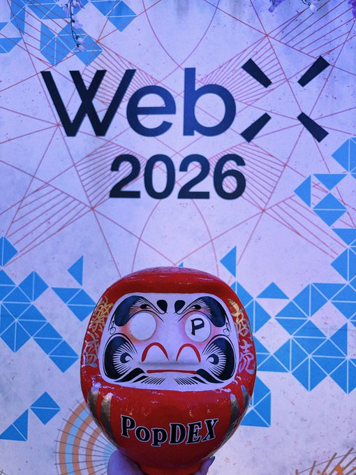 A red Daruma doll labeled "PopDEX" held up in front of a backdrop displaying "WebX 2026" with blue geometric triangle patterns.