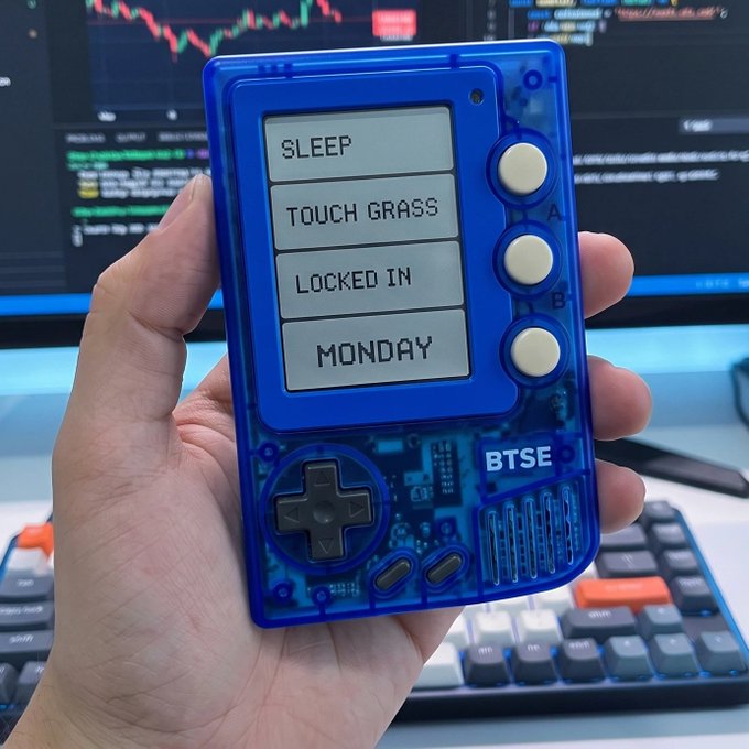 A hand holding a blue BTSE-themed retro gaming-style handheld device displaying four menu options: 'SLEEP', 'TOUCH GRASS', 'LOCKED IN', and 'MONDAY', positioned in front of a trading screen and mechanical keyboard.