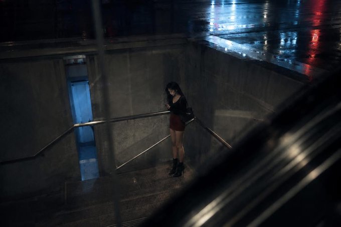 The image shows a woman standing at the top of an escalator, looking down into a dark room with a blue door. She appears to be alone and focused on her surroundings. The photo has been edited or filtered, giving it a dreamy or surreal appearance. The caption includes several words that are not directly related to the scene but could be part of the editing process.