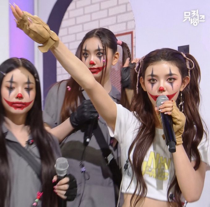 Four young women on a stage with Korean text '뮤직뱅크' (Music Bank) in the corner, wearing clown-like makeup with dark eyes and red lips; one is singing into a microphone.