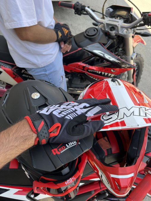 A rider holds up black and red SMK and Red Line gloves and a matching helmet in front of their red motorcycle, with another rider standing nearby in a white shirt. The image celebrates a successful day on the road, capturing the camaraderie and gear of motorcyclists enjoying a ride together.