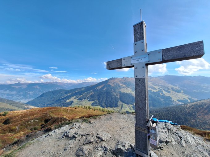 A wooden summit cross on a rocky mountain peak overlooking a vast alpine valley under a blue sky with scattered clouds.