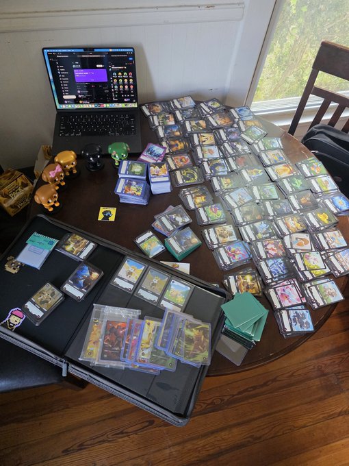A tabletop covered with hundreds of trading cards in sleeves alongside a laptop, small figurines, and an open card binder.