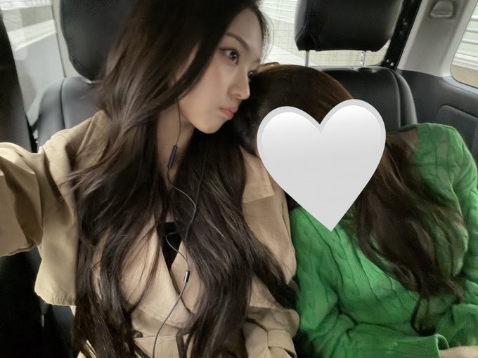 A young woman with long dark hair wearing a tan jacket takes a selfie inside a car next to a person whose face is covered by a white heart emoji, with the second person wearing a green sweater.