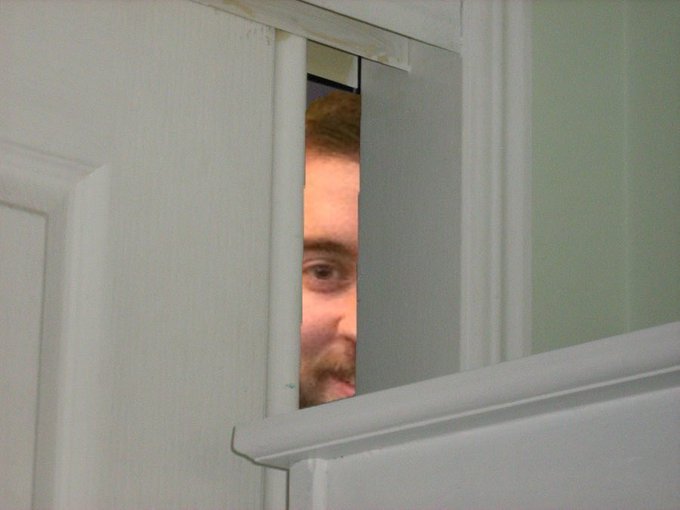 A man with a beard peeks through a narrow crack in a slightly open door, watching intently. The image humorously conveys a sense of cautious optimism or skepticism, fitting the post celebrating Chainlink's ($LINK) highest daily close against Bitcoin in over 8 months—suggesting a "peek through the door" at a potentially bullish market moment.