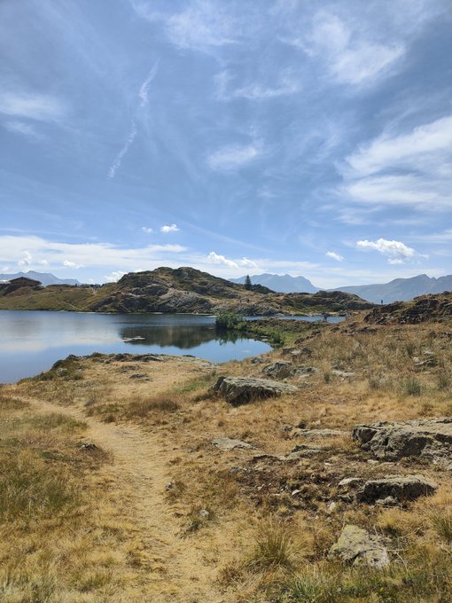 A scenic landscape photo featuring a dirt path through dry, grassy terrain leading to a calm blue lake, with rocky hills and mountains under a bright, cloudy sky.