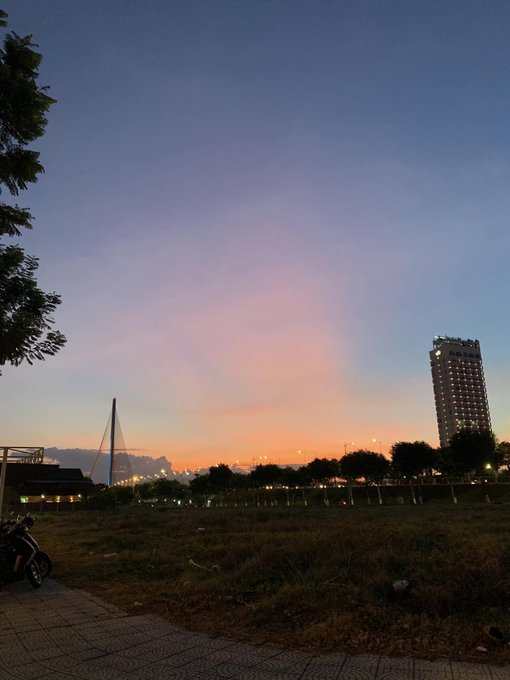 A twilight cityscape with a pink and orange sunset sky, featuring a cable-stayed bridge, a tall lit building, and a motorcycle parked in the foreground.