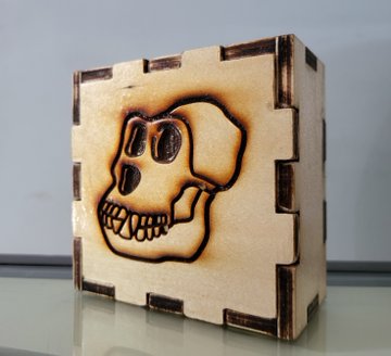 A small wooden cube box with interlocking finger joints, featuring a burned/sketched skull design on its front face, sitting on a reflective surface.