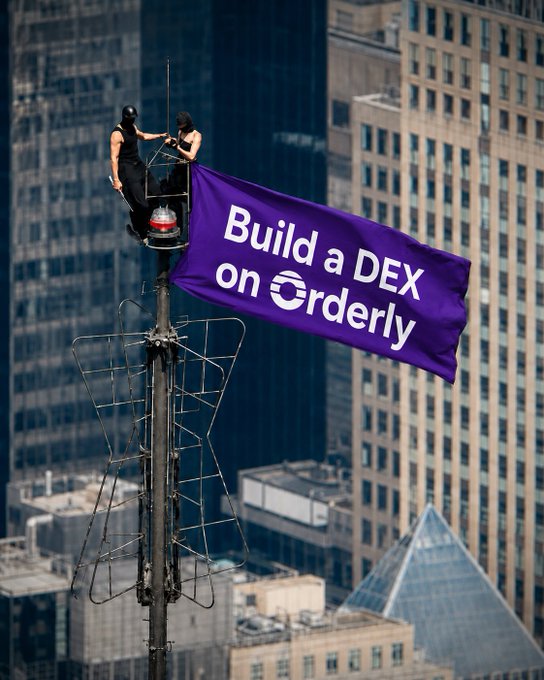 Two workers in a small elevated platform hang a purple banner reading 'Build a DEX on Orderly' from atop a communication tower with skyscrapers in the background.
