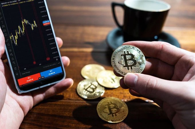 Hands holding physical Bitcoin tokens alongside a smartphone displaying a Bitcoin vs US Dollar candlestick chart on a trading platform.