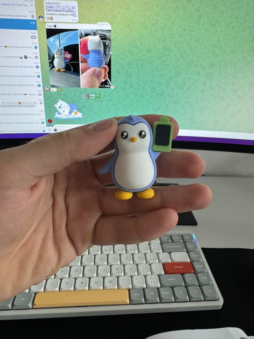 A hand holding a small blue and white penguin figurine with a green screen/keychain accessory, shown above a mechanical keyboard with a computer monitor in the background displaying images of the same figurine.