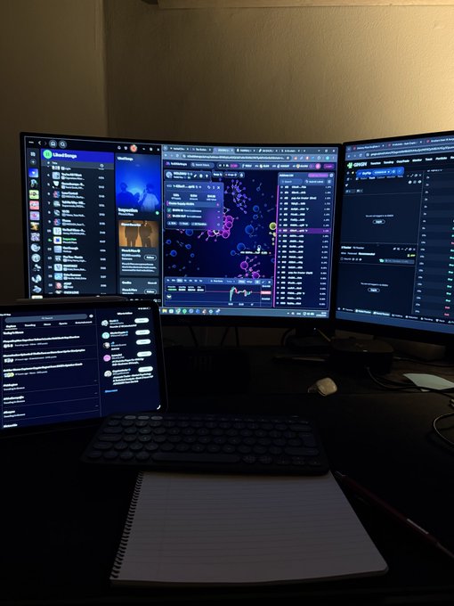 A multi-monitor trading setup in a dark room, showing crypto trading charts, Telegram chats, and messaging apps across four screens, with a keyboard and notebook on the desk.