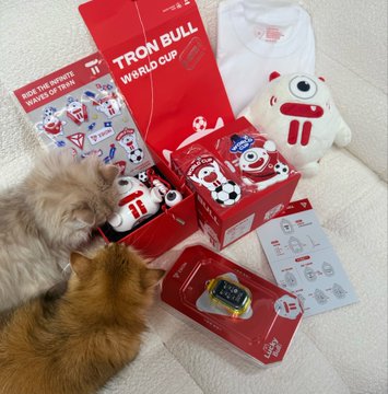 A fluffy cat lounges beside a "TRON BULL WORLD CUP" promotional display featuring Monsters Inc.-style mascot plushies, a wristwatch, and a red draw-lots (fortune-telling) device, tying into the post's playful comparison between the Tron Bull mascot and the iconic Pixar characters.