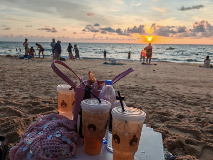 Beach scene at sunset with several cups of iced drinks and a water bottle in the foreground on a table, while people gather near the water in the background.