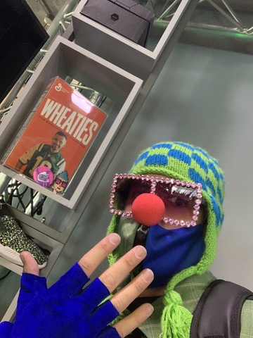 A person wearing a green and blue knit hat with a red clown nose, large pink-rhinestone sunglasses, and blue fingerless gloves poses in front of a box of Wheaties cereal featuring a man on the cover.